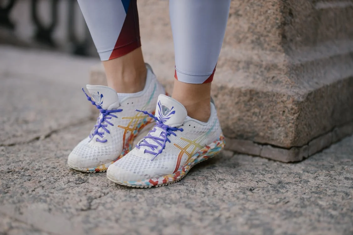 Someone wearing a pair of colorful, painted shoes and leggings stands in front of a pillar on cement.