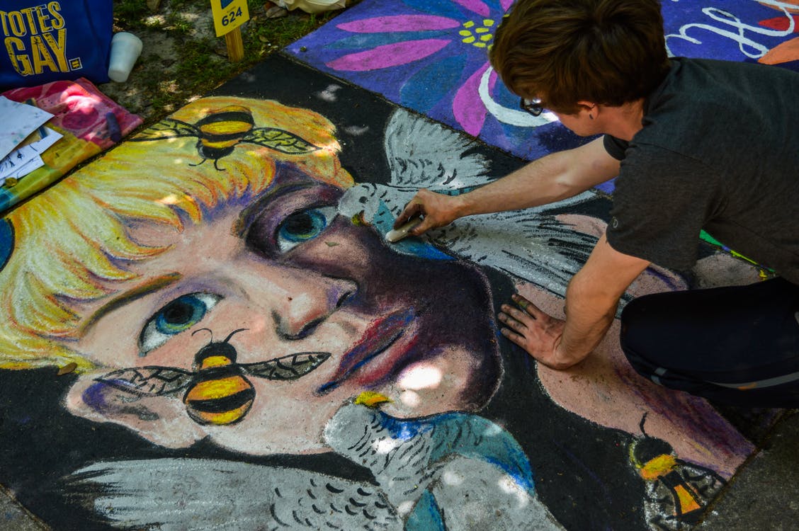 A boy is kneeling over chalk art with a piece of chalk in his hand. The artwork is of a blonde-haired boy's head surrounded by darnkess. There are 2 pigeons and 2 bees flying in front of his face.
