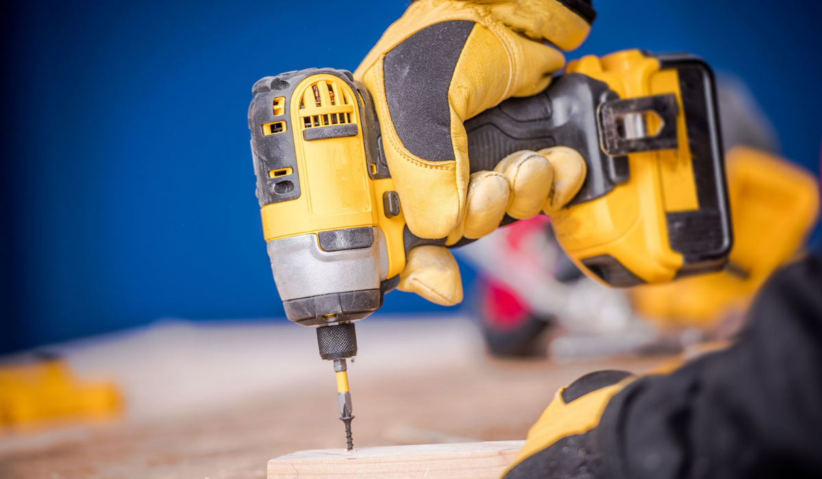 A cordless dril is being used to screw a nail into a wooden plank on a table.