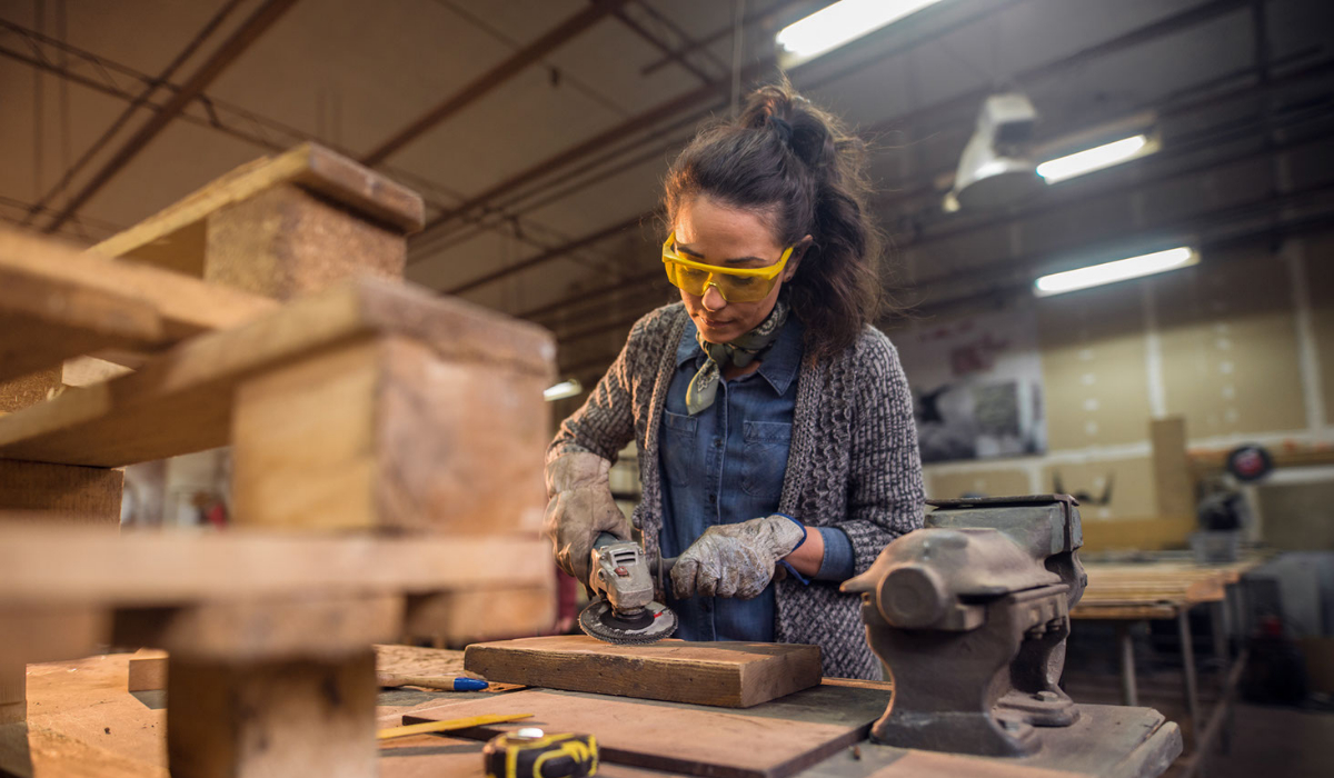 A woman wearing protective glasses and gloves is sanding down wood